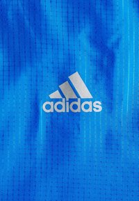 Niebieska tkanina z wzorem kratki, na której znajduje się białe logo Adidas składające się z trzech ukośnych pasków nad nazwą marki.