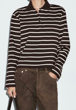 Osoba ubrana w czarno-białą koszulę polo z długim rękawem w paski, wpuszczoną w brązowe zamszowe spodnie, z jedną ręką w kieszeni.