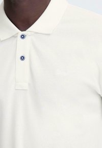 Polo crème en tissu texturé. Il présente un col classique, deux boutons bleu foncé et un logo discret sur la poitrine.