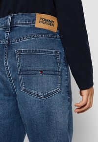 Jeans in denim blu con una toppa in pelle marrone chiaro etichettata "TOMMY HILFIGER" sul retro. Presentano due tasche posteriori e una texture di lavaggio sbiadito.