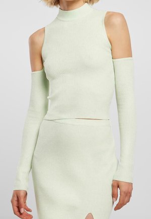 Vrouw die een lichtgroene, geribbelde, gebreide mouwloze top draagt met cold shoulder-uitgesneden details en een bijpassende taille-hoogte rok met een split aan de voorkant.