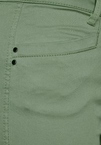 Gröna bomullsblandade byxor med en slät yta, framficka och två dekorativa svarta metallknappar ovanför fickan.
