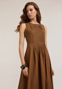 Brown sleeveless dress with vertical pleats at the bodice, flared skirt, cotton fabric, and a smooth texture. Accessorized with bracelets.