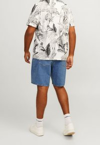 Bílé tričko s krátkým rukávem a černými tropickými listovými vzory, doplněné modrými džínovými šortkami. Outfit je doplněn bílými teniskami a žebrovanými bílými ponožkami.