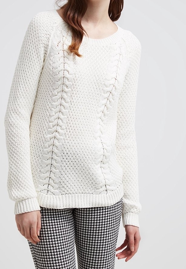 Femme portant un pull en maille blanc avec des motifs torsadés et un pantalon pied-de-poule noir et blanc sur un fond clair uni.