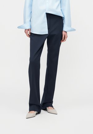 Femme portant un pantalon large bleu marine, une chemise boutonnée bleu clair, et des talons pointus beige clair, se tenant contre un fond uni.
