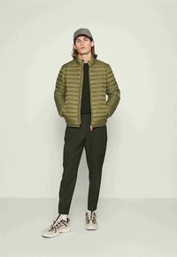 Junger Mann in einer olivgrünen Steppjacke, schwarzem Shirt, dunkler Hose, beigen Turnschuhen und einer grauen Mütze, der mit den Händen in den Taschen dasteht.