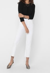 Weiße Jeans mit einem verkürzten Schnitt und fransigen Säumen, kombiniert mit einem schwarzen Langarmoberteil. Die Schuhe haben ein Schlangenhautmuster und spitze Zehen.