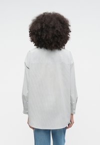 Πουκάμισο με ρίγες σε γκρι και λευκό, με μακριά μανίκια και στρογγυλεμένη τελειώματα, κατασκευασμένο από ελαφρύ ύφασμα. Ορατές ραφές και λεπτομέρειες με κουμπιά.