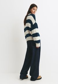 Giovane donna con lunghi capelli scuri, che indossa un maglione lavorato a maglia blu e bianco a righe, jeans larghi scuri e stivali color cammello, in piedi di lato.