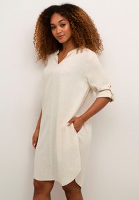 Abito in lino beige con colletto, maniche corte arrotolate, tasche laterali e orlo curvo. Tessuto testurizzato con sottili variazioni di colore.