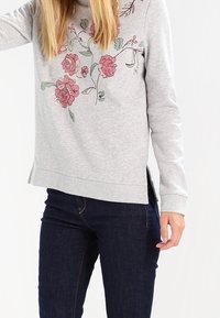Sudadera gris con diseño floral en tonos rosa y verde. Cuenta con mangas largas, puños acanalados, aberturas laterales y un corte relajado. Combinada con jeans oscuros.