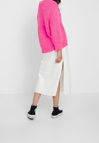 Pull en tricot rose vif, jupe en denim blanche avec fente sur le côté, et baskets montantes noires. Accent sur la couleur vive et le tissu texturé.
