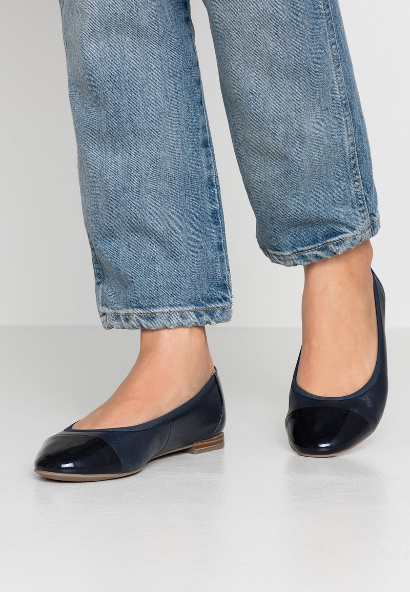 Pieds portant des ballerines en cuir bleu foncé avec des embouts brillants et un jean bleu clair retroussé sur un sol gris.