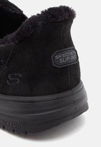 Fekete slip-on cipő, velúr felső résszel, műszőrme gallérral és gumitalppal. A hátsó részen található egy "Skechers" logó címke.
