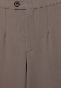 Gros plan sur un pantalon habillé taupe avec une ceinture boutonnée, des plis avant et une braguette zippée dissimulée.