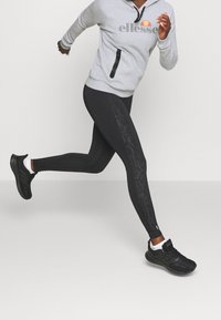 Leggings de sport noirs avec un motif texturé sur le côté, associés à un sweat à capuche gris clair avec zipper et logo, et des chaussures de course noires.