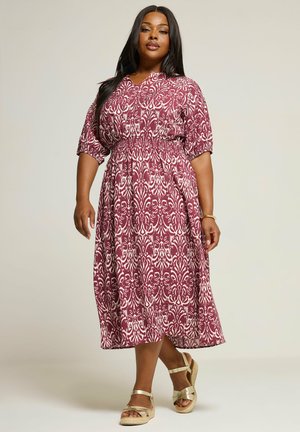 Kvinde iført en midi-kjole med bælte, mønstret i mørkerød og hvid med korte ærmer og gyldne kilehælssandaler, stående mod en ensfarvet baggrund.