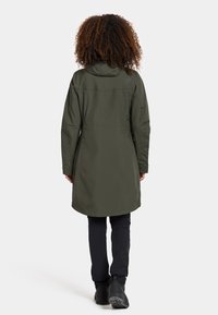 Olivgrüner wasserdichter Parka mit Kapuze, der bis zur Mitte des Oberschenkels reicht, mit geradem Schnitt und einfachem Design, kombiniert mit schwarzen Hosen und Stiefeln.