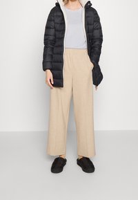 Sort pufferjakke med fleecefor, beige bukser med vide ben og sorte slip-on sko. Outfittet har en afslappet, lagdelt stil.