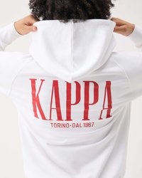 Persona con cabello rizado ajustándose una sudadera blanca con capucha que muestra un gran texto rojo "KAPPA TORINO DAL 1967" en la espalda.
