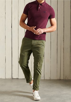 Homme portant un polo bordeaux, un pantalon cargo vert olive avec les ourlets retroussés, et des baskets blanches, debout contre un mur en bois.
