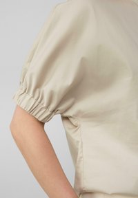 Beige Oberteil mit Puffärmeln aus glattem Material, das an den Ärmeln mit Gummizug gerafft ist und eine strukturierte, kastige Passform hat.