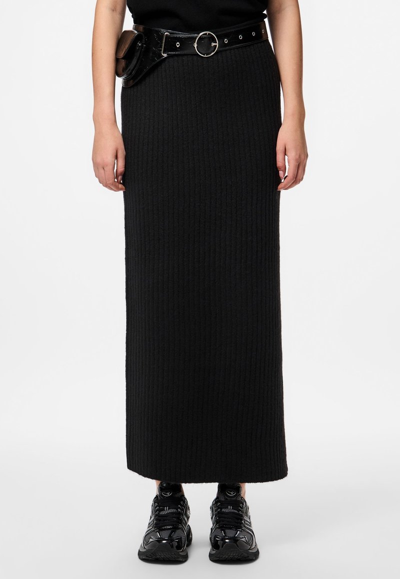 Personne portant une longue jupe noire côtelée, un sac ceinture noir et des baskets noires, debout devant un fond blanc.