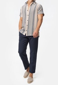 Mand iført en kortærmet beige og grå stribet skjorte med knapper, marineblå opløftede bukser og beige slip-on espadrillos, stående foran en hvid baggrund.