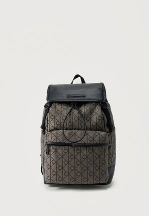 Sac à dos à motifs marron et noir avec poche avant zippée, fermeture à cordon et rabat noir sur le dessus avec poignée sur fond blanc.