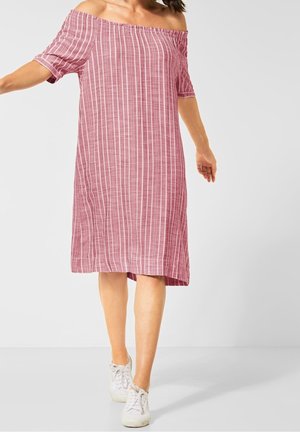 Kvinde iført knælang, off-shoulder, lyserød kjole med hvide lodrette striber, kombineret med hvide sneakers, stående på en lys baggrund.