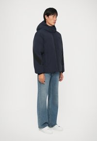 Casaco acolchoado em azul-marinho com capuz, apresentando detalhes pretos nas mangas. Combinado com jeans claros em corte fluido e sapatilhas brancas.