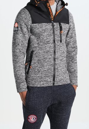 Personne portant un sweat à capuche Superdry gris zippé avec détail capuche noir et un pantalon de jogging foncé assorti avec des patchs Superdry, mains dans les poches.