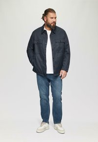 Dunkelblaue Knopfjacke mit zwei Vordertaschen, getragen über einem weißen T-Shirt, kombiniert mit blauen Jeans und hellen Sneakers.