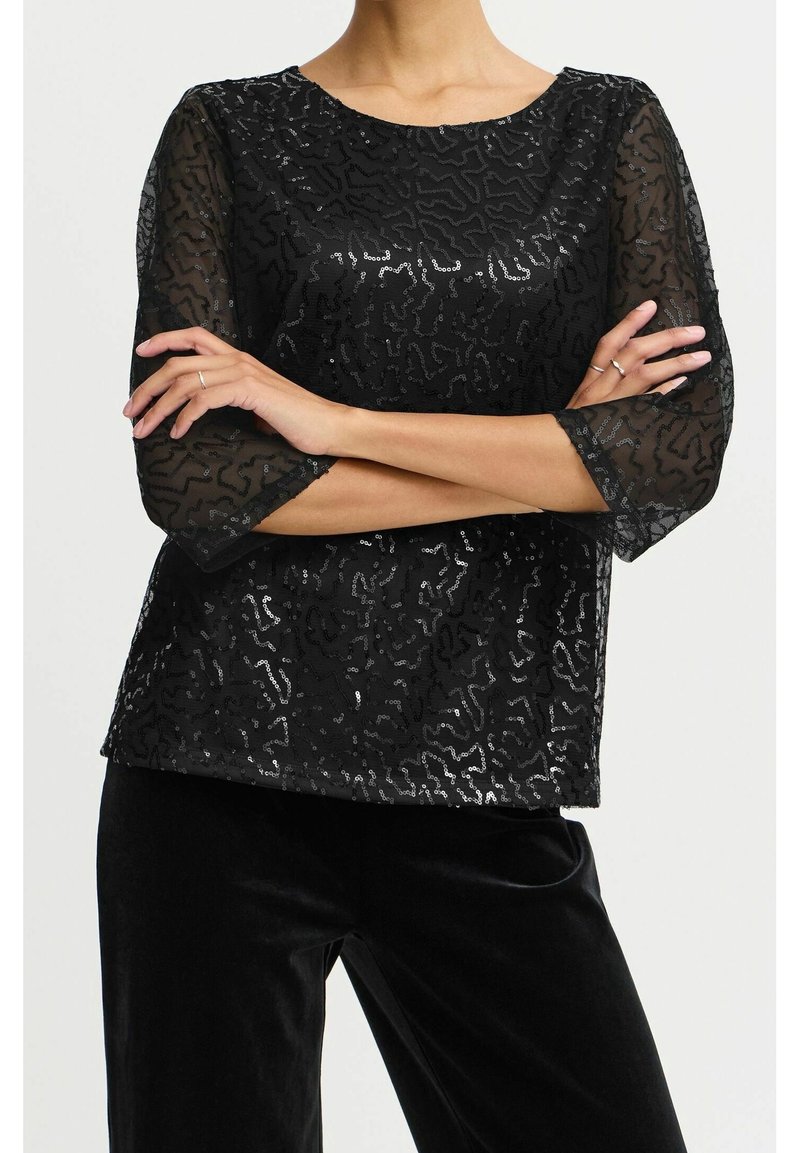 Haut noir avec des manches transparentes texturées arborant un motif ondulé de sequins. Encolure arrondie, coupe décontractée, associé à un pantalon en velours noir.