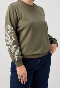 Felpa verde oliva con ricamo floreale bianco sulla manica sinistra, polsini e orlo a costine, e scollo rotondo, abbinata a jeans blu.