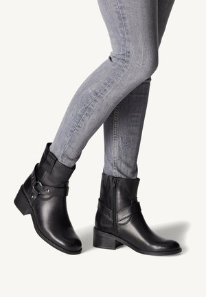 Black leather ankle boots with side zip, adjustable buckle accents, rounded toe, and a low heel, paired with gray skinny jeans.