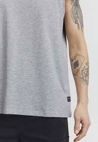 Graues ärmelloses Shirt aus strukturiertem Stoff, versehen mit einem schwarzen Etikett am Saum. Der Arm ist mit komplizierten Tattoos und einem Motiv von Spielkarten verziert.