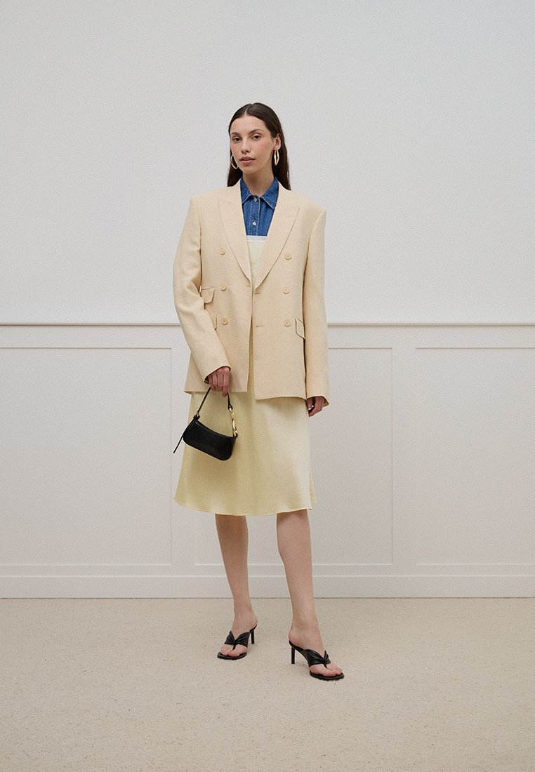 Beige, doppelt geknöpftes Blazer über einem Jeanshemd, kombiniert mit einem blassen gelben Kleid. Eine schwarze Handtasche und schwarze, hochhackige Sandalen runden den Look ab.