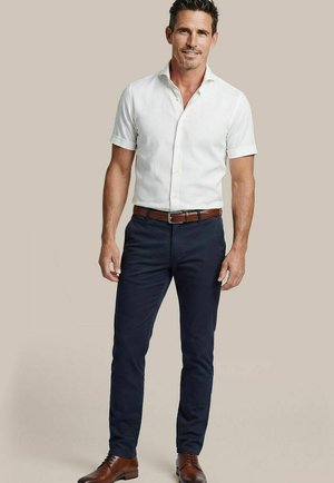 Mann trägt ein kurzärmeliges weißes Hemd mit Knöpfen, marineblaue Hose, braunen Gürtel und braune Lederschuhe, steht vor einem einfarbigen beigen Hintergrund.