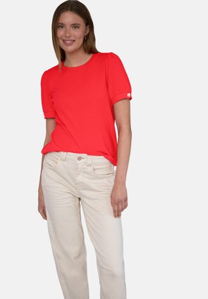 Femme souriante portant un chemisier rouge à manches courtes et un pantalon beige clair sur un fond blanc uni.