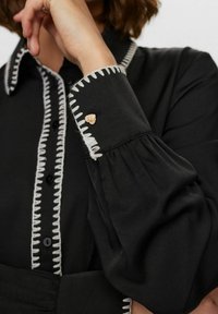 Black shirt featuring a white embroidered edge along the collar and cuffs, with a decorative cream heart-shaped button on the sleeve.