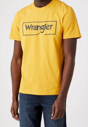 Homme portant un t-shirt jaune à manches courtes avec le logo noir "Wrangler" et un jean bleu, debout devant un fond clair uni.
