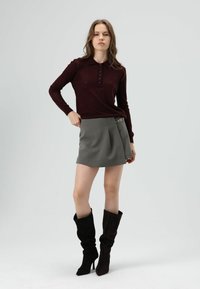Polo en tricot bordeaux à manches longues avec motif en losanges, associé à une mini-jupe plissée grise ornée d'un détail chaîne sur le côté, et des bottes noires montantes jusqu'aux genoux.