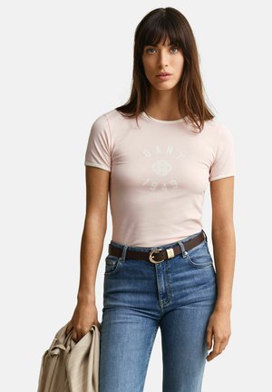 Vrouw met lang bruin haar die een lichtroze GANT 1949 T-shirt draagt, gestopt in een blauwe spijkerbroek, en een beige jas vasthoudt in haar linkerhand.