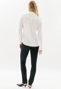 Camisa blanca de manga larga con textura suave, dobladillo curvado y puños abotonados, combinada con pantalones ajustados negros y zapatos de tacón negros.