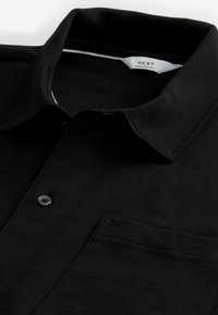 Musta poolopaita, joka on valmistettu pehmeästä kankaasta. Paidassa on kaulus, yksittäinen napitus ja vasemmalla puolella rintatasku. Tagi näkyvissä kauluksessa.