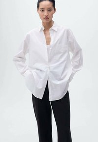 Camicia bianca over-size in cotone crudo, con chiusura a bottoni, colletto, maniche lunghe e taschino sul petto. Abbinata a pantaloni neri.