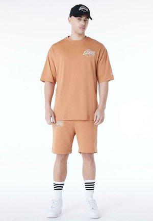 Man wearing tan Los Angeles Lakers T-shirt and matching shorts, black Lakers cap, white striped socks, and white sneakers standing facing forward.