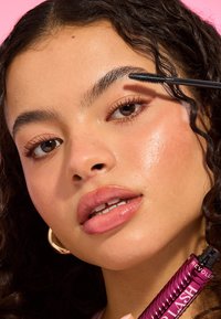 Maskaraty tube i rosa metallisk innpakning som holdes nær ansiktet. Modellen har glansfulle lepper, definerte øyenbryn og subtil øyenmakeup mot en rosa bakgrunn.